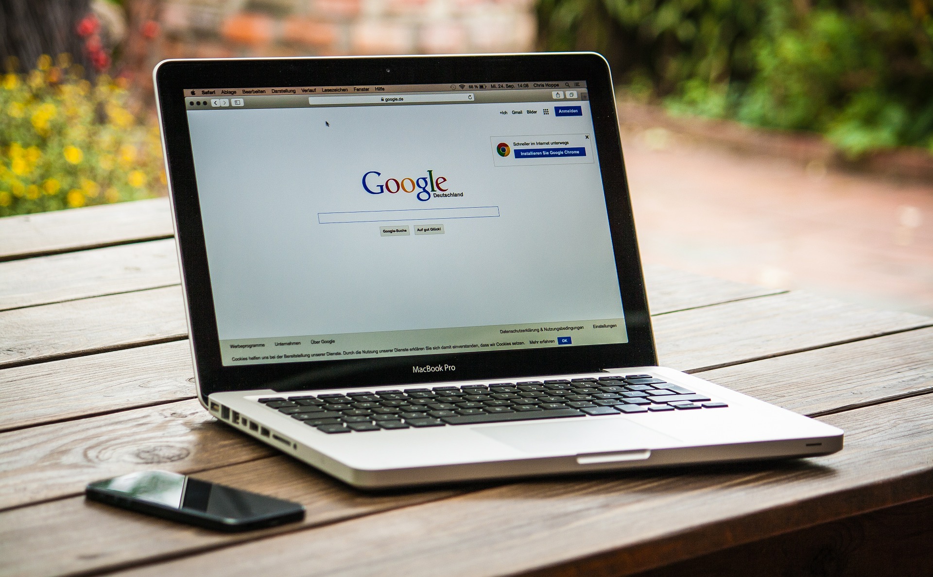 Photo of an open laptop on an outside table, screen shows the Google homepage, a smartphone is next to the laptop