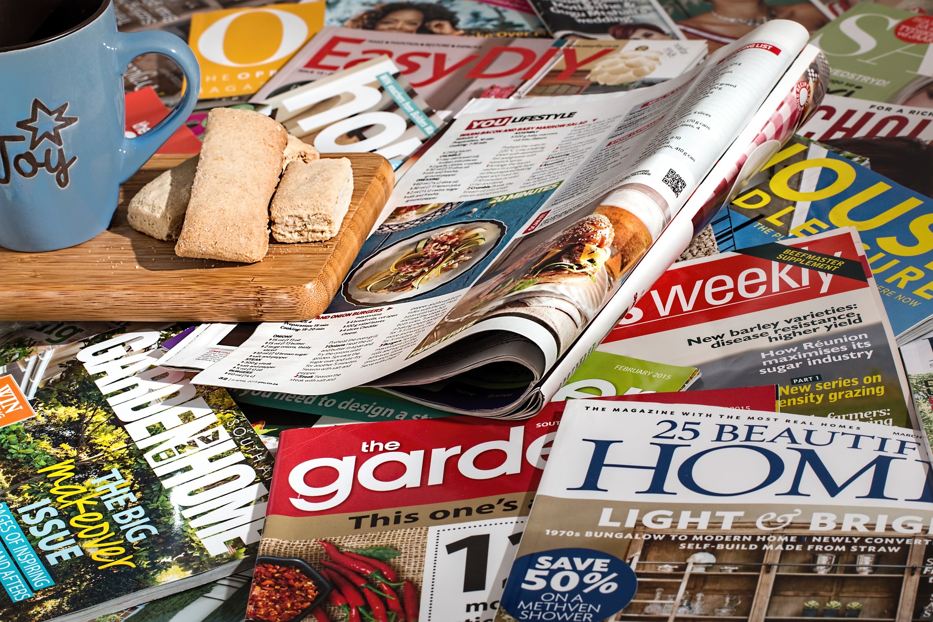 Multiple consumer magazines scattered across a tabletop. Small board with a coffee mug and bread on top of the magazines.